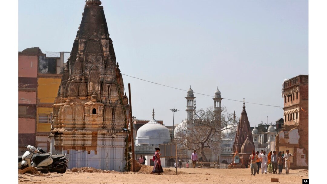 In this March 19, 2019, photo, 17th-century Gyanvapi mosque, white structure sandwiched between Hindu temples, is seen in the background as Hindu devotees walk at the site of a proposed grand promenade connecting the sacred Ganges river with a centuries-old temple dedicated to Lord Shiva, in Varanasi, India. (AP Photo/Altaf Qadri)