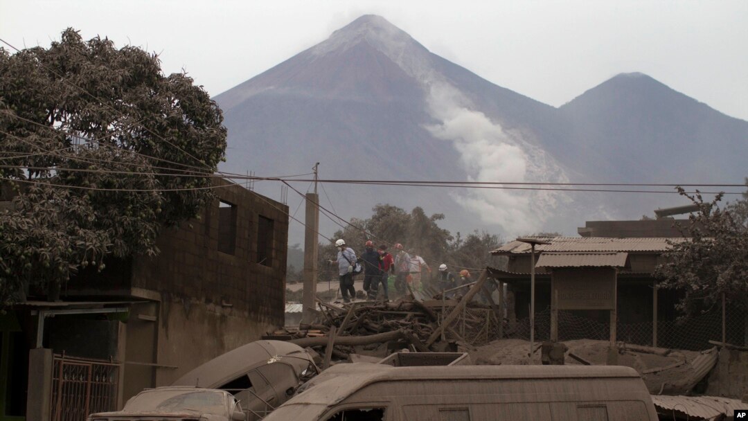 Rescue workers walk on rooftops in Escuintla, Guatemala, June 4, 2018, blanketed with heavy ash spewed by the Volcan de Fuego, or "Volcano of Fire," pictured in the background, left center. 