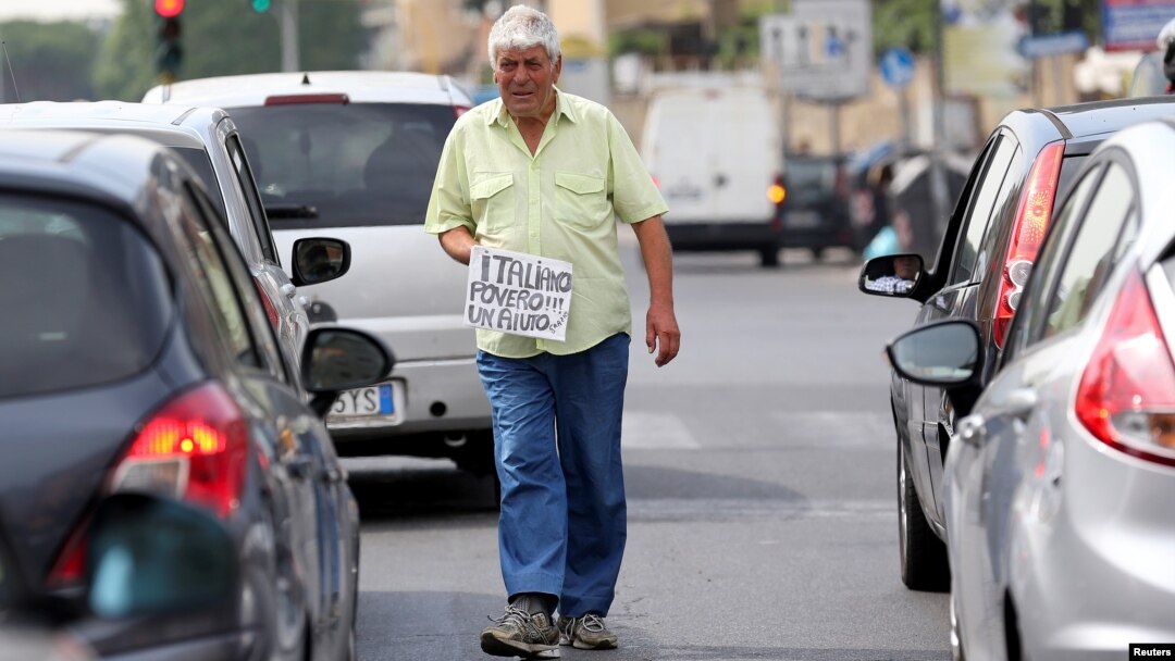 FILE - An Italian man walks between cars as he holds a banner reading "I'm a poor Italian, help me, thanks" in Rome, May 28, 2018. 