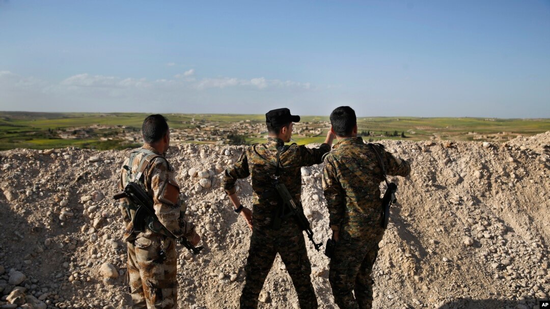 FILE -Fighters from the U.S-backed Syrian Manbij Military Council stand behind a sand barrier as they look toward a Turkish-backed fighters' position at the front line of Halawanji village, north of Manbij town, Syria, March 29, 2018. 